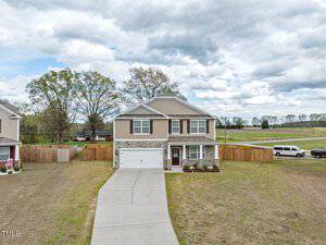 A two-story house with light brown siding and stone accents, featuring a white garage door. The front yard has a wide driveway and sparse landscaping. In the background, there are trees, open fields, and a white van parked on the side.