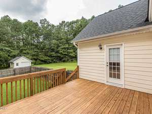 A wooden deck extends from a beige house, featuring a glass door with a light fixture above. The deck overlooks a grassy backyard surrounded by a wooden fence, with a small shed in the distance and trees lining the perimeter.