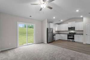 Modern open-concept interior featuring a living area with beige carpet, adjacent to a kitchen with white cabinets, stainless steel appliances, and wood flooring. A sliding glass door opens to a grassy outdoor view, and a ceiling fan is overhead.