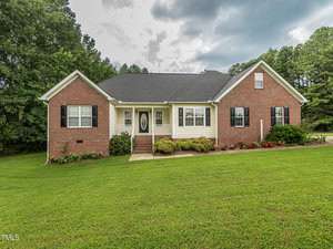 A single-story brick house at 3081 Wren Road boasts a black roof, white trim, and black shutters. The inviting front door with sidelights is framed by manicured bushes and a sprawling green lawn. Nestled in the charming community of Creedmoor, trees stand tall against a cloudy sky backdrop.