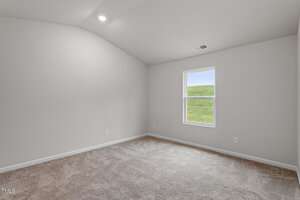 An empty room with beige carpet and light gray walls. The ceiling is vaulted, and there is a single window on one wall, displaying green grass outside. The room appears well-lit and spacious.