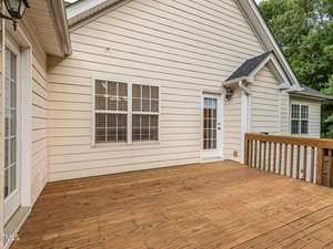 A wooden deck attached to a beige house with white-framed windows and a glass door. The deck appears spacious, surrounded by a wooden railing. The exterior has vertical siding, and trees are visible in the background.