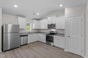 Modern kitchen with white cabinets, stainless steel appliances including a refrigerator, dishwasher, and oven. Granite countertops and light wood flooring. Single window above the sink allows natural light to brighten the space.