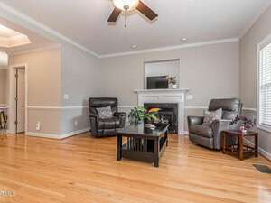 A cozy living room featuring two gray recliners, a black coffee table with plants, and a fireplace with a white mantel. The room has hardwood floors, neutral walls, and a ceiling fan. Natural light enters through a window on the right.