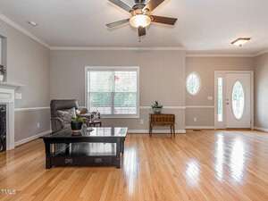 A spacious living room features light wooden flooring and neutral-colored walls. A ceiling fan with lights hangs above. The room includes a large window, a recliner, and a coffee table with plants. A decorative front door with an oval glass panel is visible.