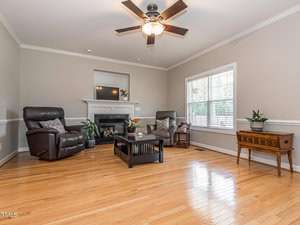 A spacious living room with light hardwood floors and neutral walls. Two recliners face a black fireplace with a white mantel. A dark coffee table is centered on a rug. A ceiling fan hangs from above, and a window lets in natural light.