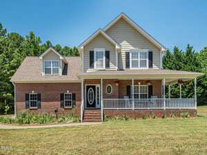 Located at 3080 Wren Road in Creedmoor, this two-story house boasts red brick and beige siding with a charming covered front porch featuring white railings. The home, adorned with multiple windows and black shutters, has a gabled roof and is surrounded by a manicured lawn with trees in the background.