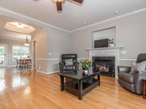 Living room with wooden floors, beige walls, and a ceiling fan. Features a black coffee table with a plant, two dark leather chairs, and a fireplace. A dining area is visible in the background.