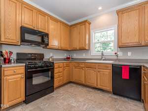A modern kitchen with wooden cabinets and tile flooring. It features a black stove and microwave, black dishwasher, and a stainless steel sink. A small window above the sink lets in natural light. A red towel hangs on the dishwasher handle.