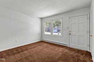 Empty room with white walls and brown carpet, featuring a window and a closed door on the right. Sunlight from the window brightens the space, and outside, a view of trees and houses is visible.