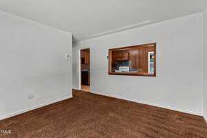 An empty room with white walls and brown carpet. A rectangular wooden-framed opening shows a view into a kitchen with brown wooden cabinets. Adjacent doorway leads to another part of the house.