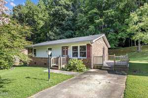 A small, single-story brick house with a front porch and a wooden ramp. It is surrounded by lush green grass and tall trees. A driveway and a lamppost are in the foreground.