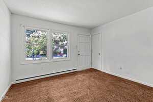 A small, empty room with white walls and a brown carpet. There are two windows showing a view of greenery outside, a white door, and a closed door leading to a closet. The room is well-lit with natural light.