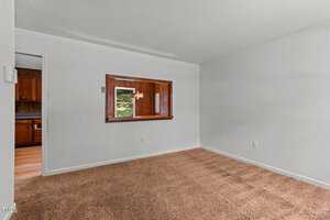 Empty room with brown carpet and white walls. A wooden-framed window opens to a view of a kitchen with wood cabinets and visible lighting. Doorway leads to adjacent space with wood flooring and kitchen.