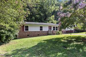 A small brick and white-paneled house with a sloped roof sits surrounded by lush greenery. Blooming trees frame the yard, and a wooden porch is visible at the entrance. The grass lawn in front is well-maintained and sunlit.
