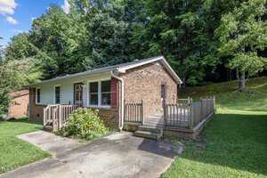 A single-story brick house with a wooden ramp leading to the front door. It's surrounded by a lush green yard and trees. The sky is clear and blue, indicating a sunny day.