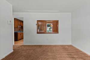 An empty room with white walls and brown carpet. There is a large open window looking into a wood-paneled kitchen area with dark cabinets and a glimpse of a stove and sink. Natural light filters in through a window.