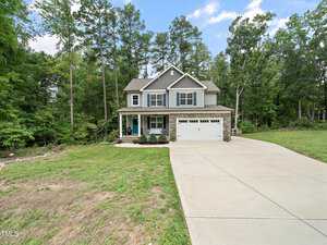 Nestled among tall green trees, the two-story gray house at 2467 Golden Forest Drive in Franklinton boasts a welcoming porch. It features a two-car garage and a wide concrete driveway, while the lawn, partially grass-covered, extends to the edge of a wooded area under a blue sky.