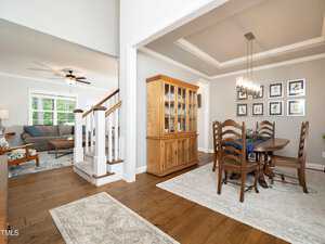 A bright, open-concept living and dining area with hardwood floors. The dining room features a wooden table with chairs and a matching cabinet. The living room has a gray sofa, armchair, and a ceiling fan. Artwork adorns the walls.
