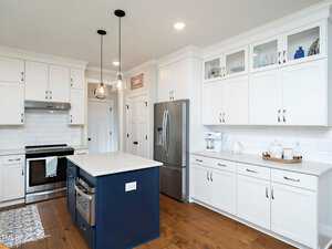 A modern kitchen with white cabinets, a stainless steel refrigerator, and a dark blue island with a white countertop. Pendant lights hang above the island, and a patterned rug lies on the wooden floor.