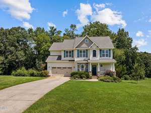 A two-story suburban house with a gray roof and beige siding sits at the end of a concrete driveway. It is surrounded by a well-manicured lawn and trees. The sky is blue with scattered clouds.