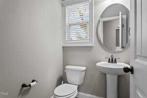 A small, neatly arranged bathroom with light gray walls. It features a white toilet, an oval mirror above a pedestal sink, and a window with horizontal blinds letting in natural light. A toilet paper holder is mounted on the wall.