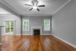 A spacious living room with light green walls, hardwood flooring, and a white fireplace centered between two windows. A ceiling fan with lights is above, and a doorway leads to a wooden deck outside.