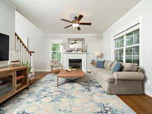 A modern living room with a gray sectional sofa, blue patterned rug, and wooden coffee table. A fireplace with a mirror above it is centered on the wall. Ceiling fan and large windows provide light. A staircase is visible to the left.