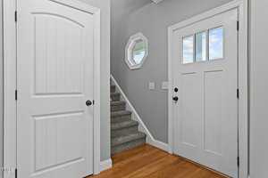 A hallway with a light green wall, white interior door on the left, a white front door with windows on the right, and a carpeted staircase leading upward. A small wall mirror with a hexagonal frame is mounted above a light switch.