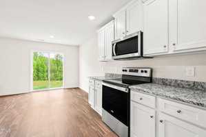 Bright modern kitchen with white cabinets, granite countertops, and stainless steel appliances. Natural light fills the room from a large sliding glass door, leading to a backyard with green foliage and a wooden floor extending throughout the space.