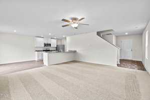 Spacious open-plan living area featuring a carpeted floor, ceiling fan, and a view of the modern kitchen with stainless steel appliances and white cabinetry. A staircase leads to the upper floor, and the walls are painted in a soft, light color.