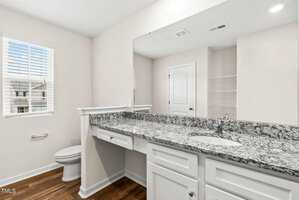 A bright, modern bathroom featuring a large granite countertop with a sink, white cabinetry, and a toilet. A window with blinds is on the left, allowing natural light to fill the space. A door and a built-in shelving unit are visible in the background.