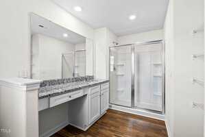 Modern bathroom with a large mirror above a granite countertop, white cabinets, and a glass-enclosed shower. The floor is wooden, and the walls are painted light beige. Shelving is visible to the right.