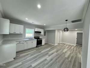 A modern kitchen and living area with light wood flooring. The kitchen features white cabinets, a dark stove, and a window above the sink. Recessed lighting and a spherical pendant light illuminate the open space.