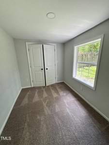 A small, gray-carpeted room with light gray walls. There is a window on the right, providing natural light, and a closed white double-door closet at the end of the room. A fenced yard is visible through the window.