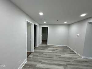 A modern, empty room with light gray walls and wood-look flooring. The room has recessed lighting on a white ceiling and two doorways, one leading to a hallway. There's a thermostat mounted on the wall.