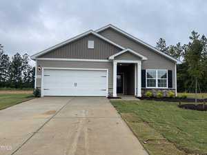 A single-story house on 201 Shelfit Trail features gray siding, white trim, and a two-car garage. The Oxford-style driveway and newly planted lawn lead to a covered entrance, with trees and clouds in the background.