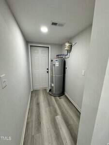 A narrow, white hallway with a wooden floor leads to a closed white door. A tall, cylindrical water heater with attached pipes is situated against the right wall. Ceiling lights illuminate the space.