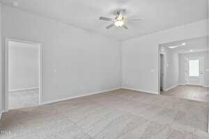 A bright, empty room with beige carpet and white walls. A ceiling fan is centered on the ceiling. Two open doorways lead to adjacent rooms, and a hallway with wooden flooring leads to a white door at the back. Natural light filters in.