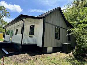 A small, single-story house at 217 Clement Avenue boasts a charming front porch with white and gray siding beneath its black roof. Nestled in Oxford, it is surrounded by lush trees and grass. The clear, sunny sky complements the view, and an air conditioning unit rests subtly to the side.