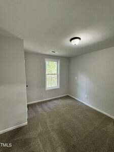 A small empty room with gray walls and a beige carpet. There is a single window with a view of greenery outside. A ceiling light fixture is visible, and a partially open door is on the left side of the image.
