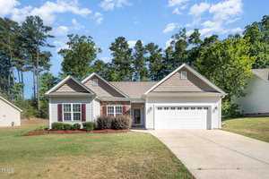 A single-story house with a light-colored facade and a two-car garage is surrounded by a well-maintained lawn. The background features tall trees and a blue sky with scattered clouds.