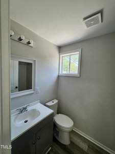 A small bathroom with a sink and cabinet on the left, a mirror above it, and a white toilet on the right. A window with natural light is on the upper right wall. The floor has light wood-like tiles.