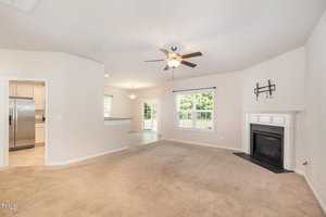 Spacious living room with beige carpet, white walls, and a ceiling fan. A fireplace is in the corner, and large windows let in natural light. The room leads to a bright kitchen with stainless steel appliances and a dining area.