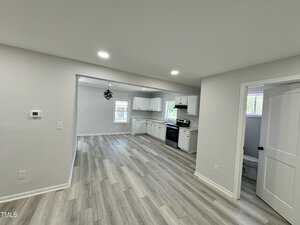 A modern kitchen and dining area in a home, featuring light gray walls, laminate wood flooring, white cabinets, a black stove, and a hanging light fixture. A doorway leads to a small bathroom. Natural light fills the space through the windows.
