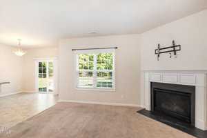 Living room with beige carpet, a white fireplace, and wall mount for a TV. Large window with blinds overlooks a grassy yard. Adjacent dining area with tiled floor and hanging light fixture. Sliding glass door leads to an outdoor space.