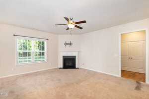 A spacious, empty living room with beige walls and carpet, featuring a white fireplace with a TV shelf above. A ceiling fan with lights hangs from the ceiling. Large window with blinds on the left lets in natural light. Doorway visible on the right.