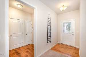 A bright hallway with a wooden floor, white walls, and two closed white doors on the left. A decorative ladder with framed photos leans against the wall. A door with patterned glass is at the end, under a ceiling light fixture.