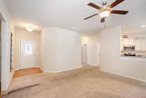 A spacious, empty room with beige carpet and light-colored walls. It features a ceiling fan with lights, a kitchen area with stainless steel appliances in the background, and a white front door. Natural light illuminates the space.