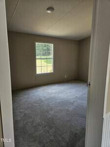 An empty room with light gray carpet and beige walls. A window on one wall offers a view of a grassy area and trees outside. The ceiling has a small round light fixture.